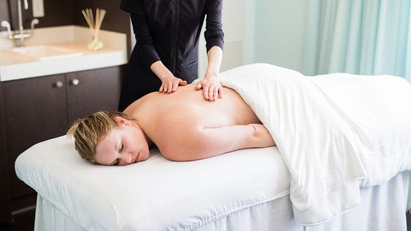 A woman receives a massage on a treatment table, relaxed under a white sheet, while a therapist works on her back.