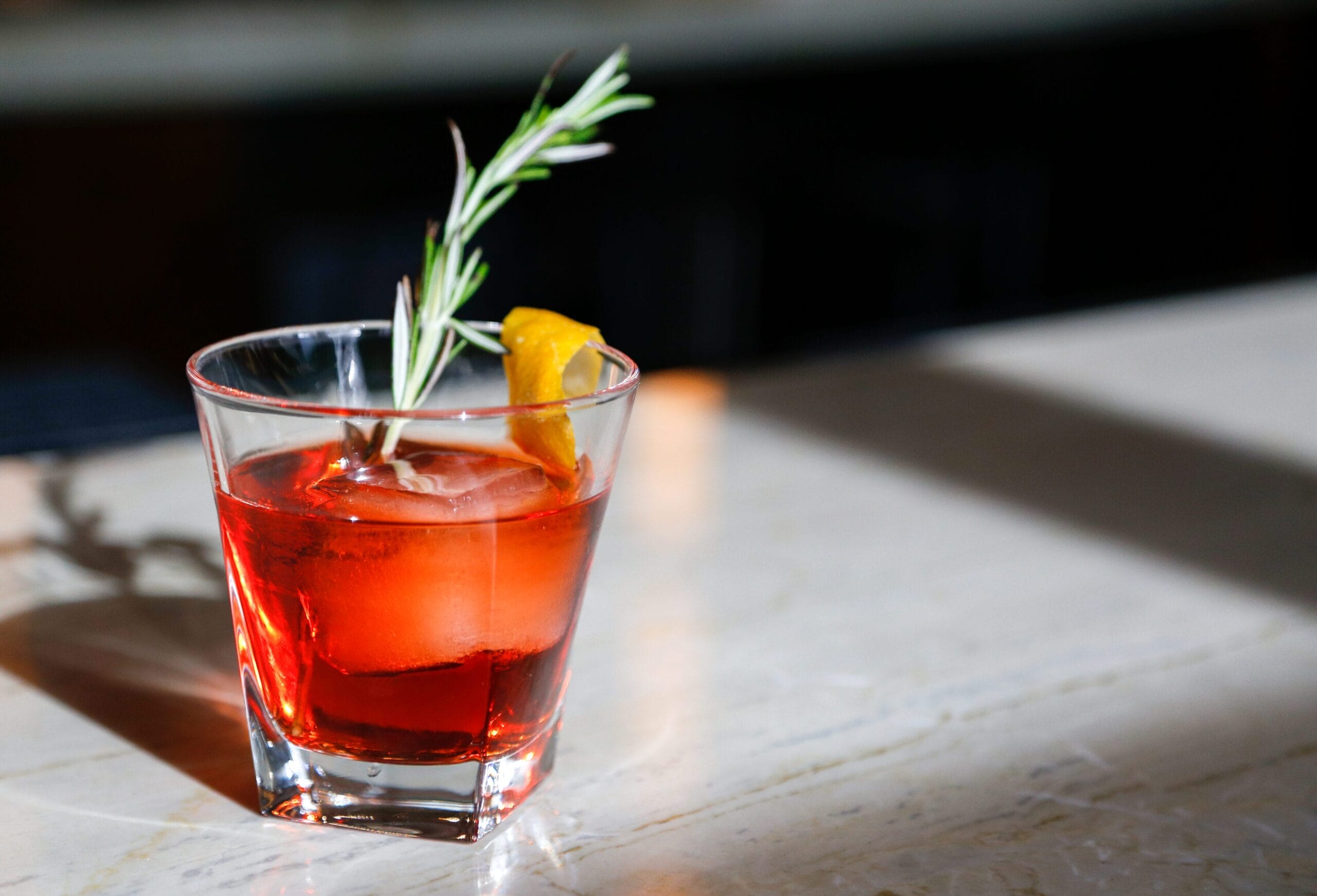 A glass of vibrant red cocktail garnished with a lemon twist and a sprig of rosemary on a marble surface.