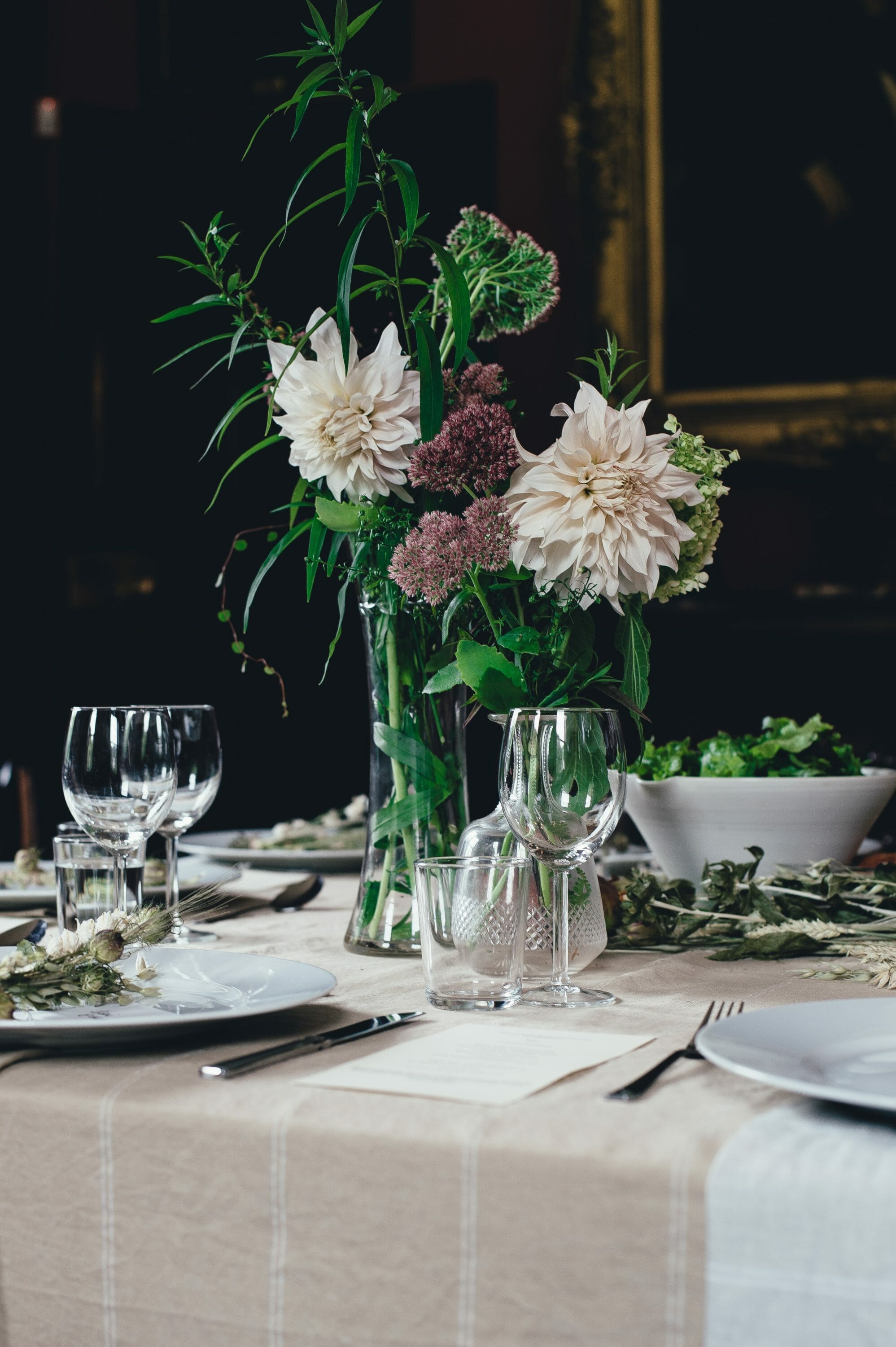 Elegant dining table set with a floral centerpiece, glassware, and a bowl of salad, against a dark, rich backdrop.