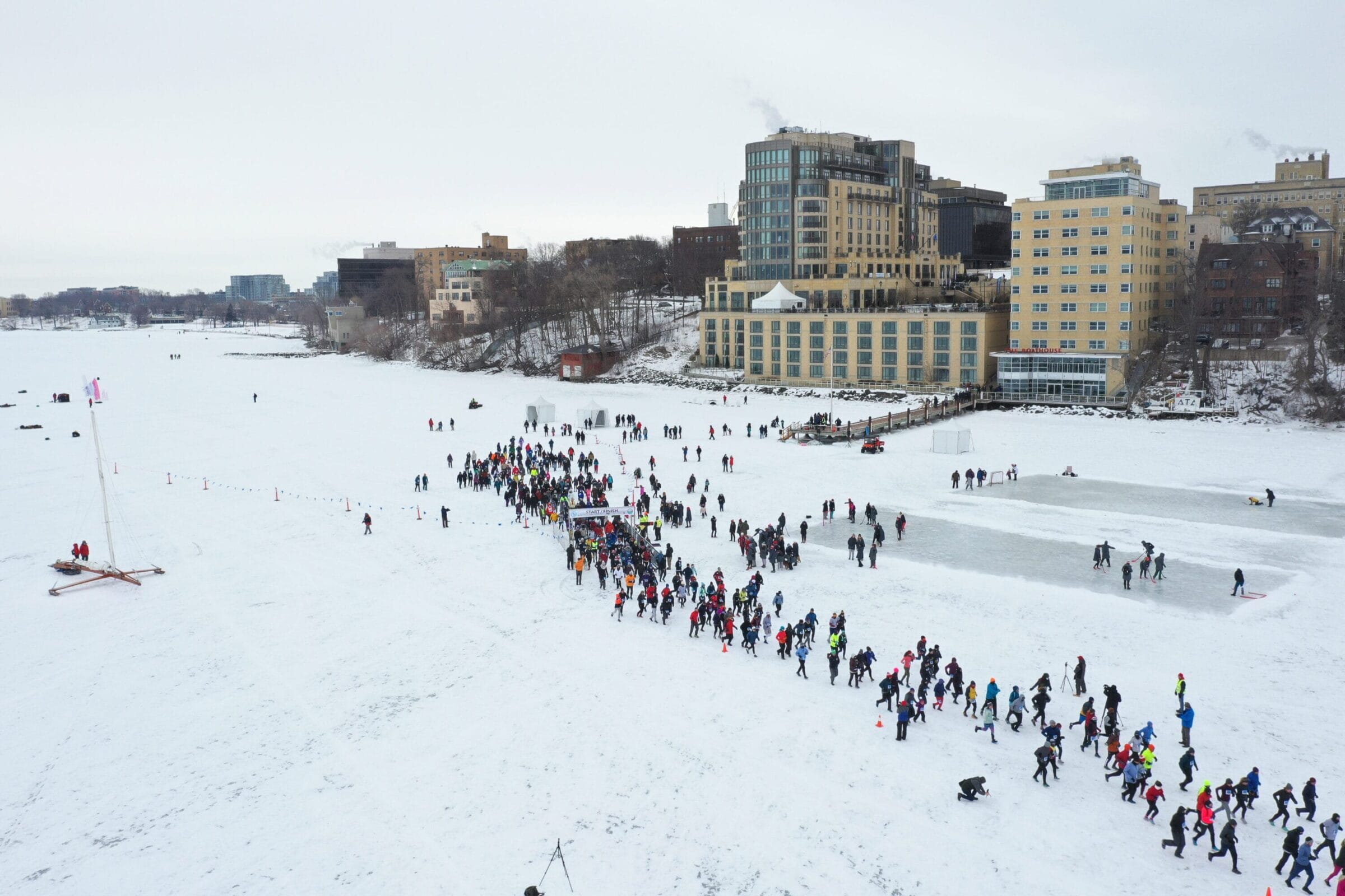 Crowd of people walking across a snowy landscape near a frozen lake, with buildings and a sailboat in the background.