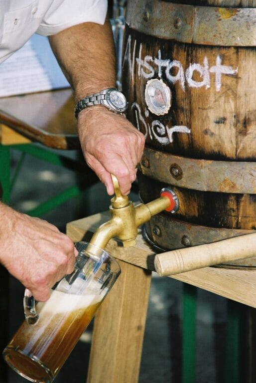 A hand pours beer from a brass tap into a glass, with a wooden barrel labeled "Altstadt Bier" in the background.