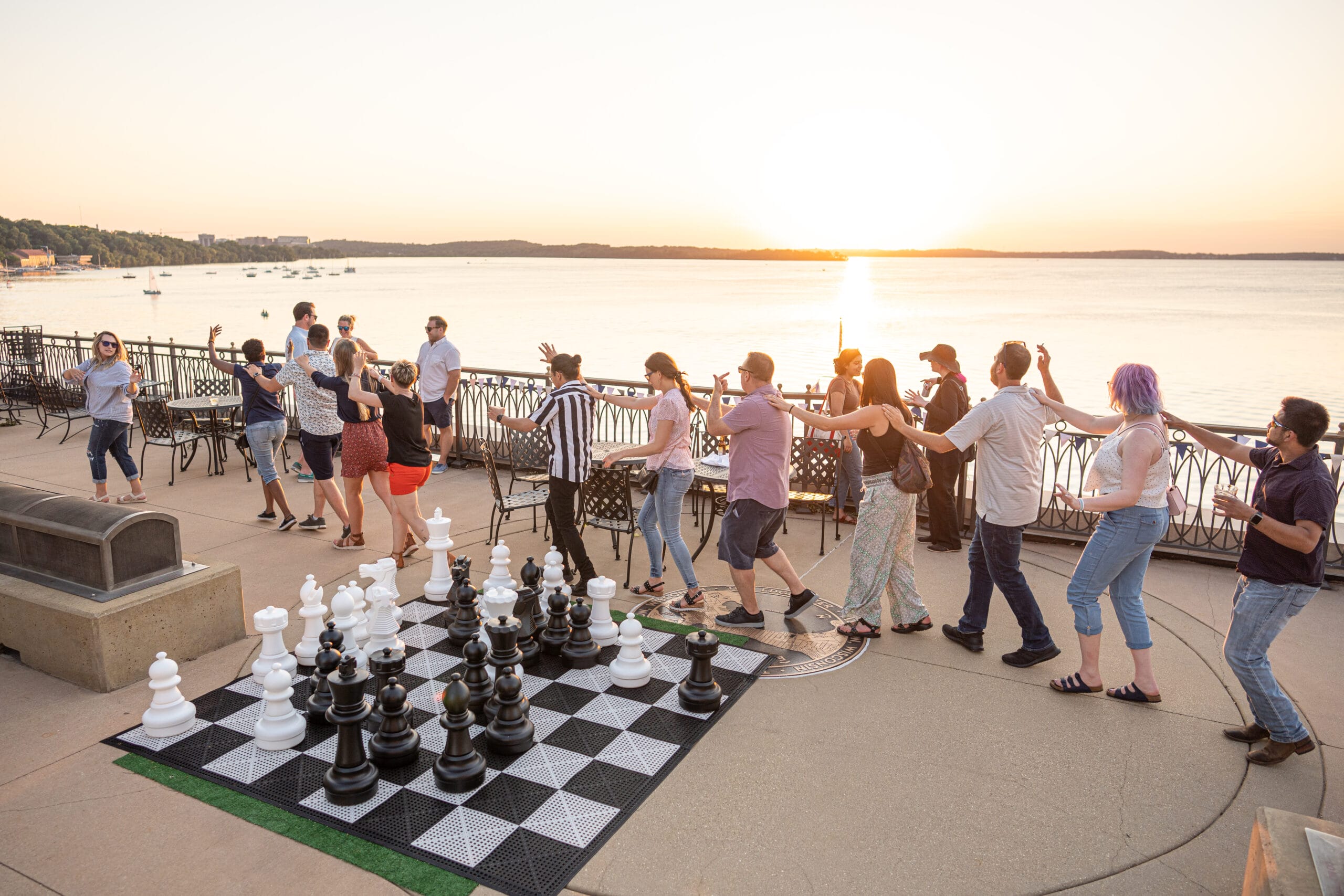 A group of people dance near a large outdoor chess set at sunset by the water, with chairs and boats in the background.