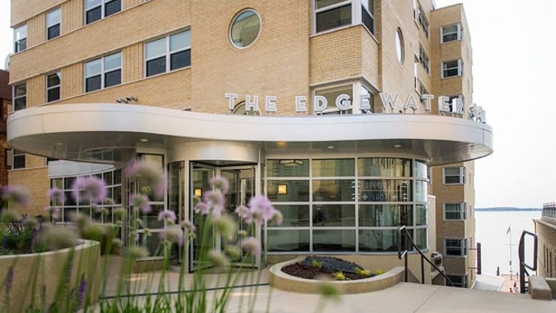 Modern building entrance of The Edgewater, featuring large glass doors and flowering plants in the foreground.