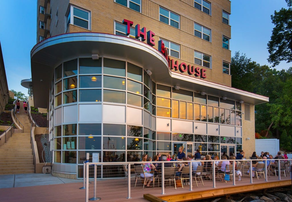 Modern restaurant with large glass windows, outdoor seating, and a staircase leading up, surrounded by greenery.