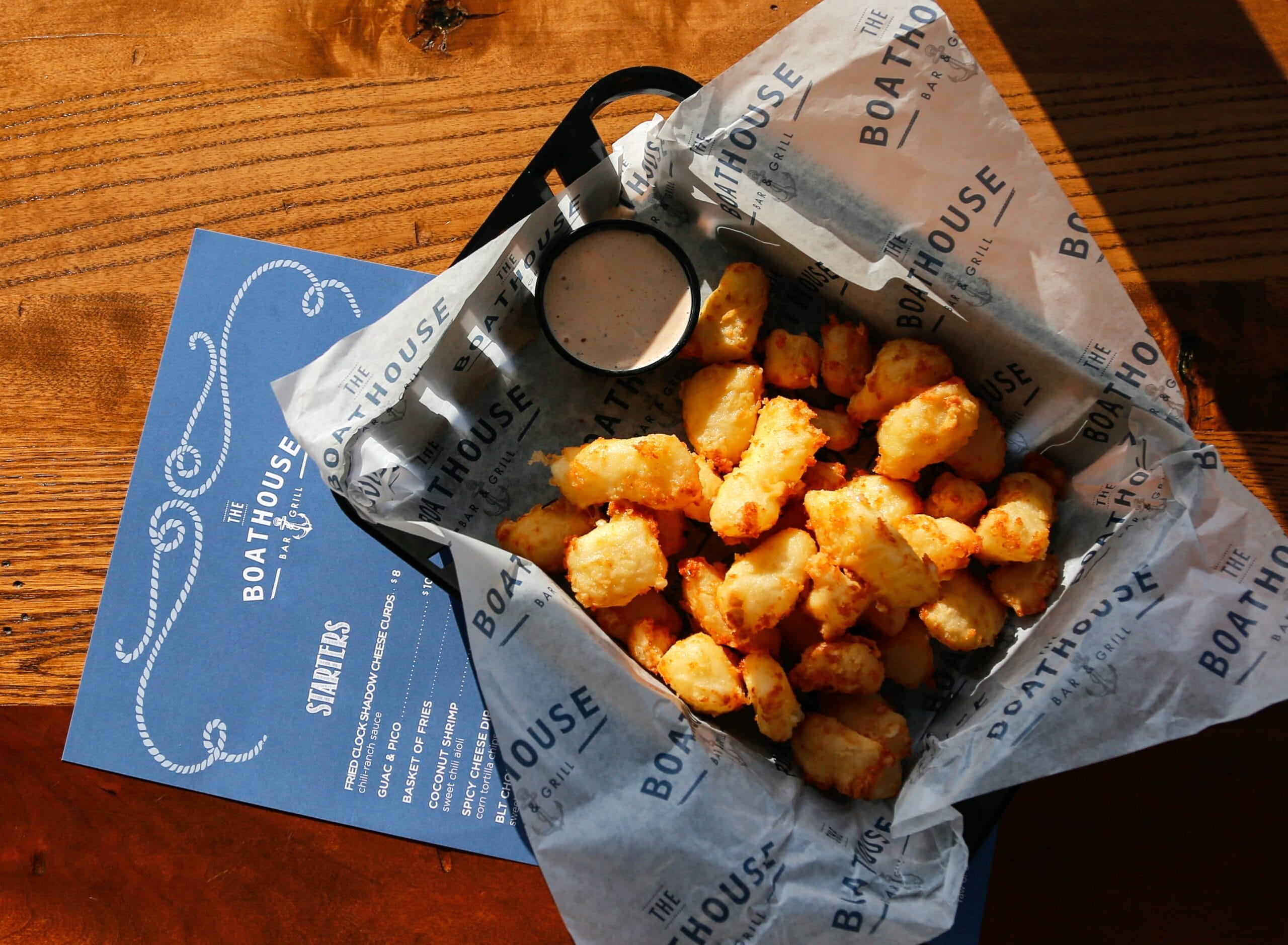 Golden, crispy cheese curds in a basket with a side of dipping sauce, resting on a wooden table next to a menu.