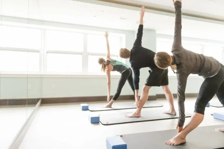 Three individuals practicing yoga in a bright studio, stretching towards the ceiling with yoga mats and blocks.