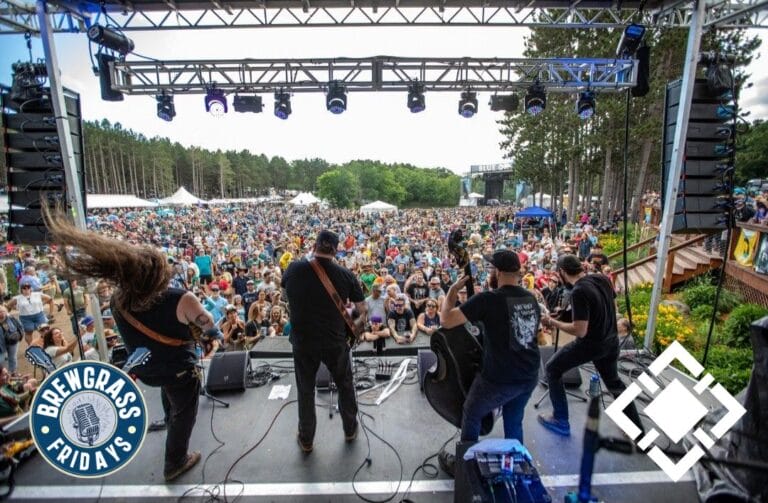 Musicians perform on stage before a large crowd at an outdoor festival, surrounded by trees and tents.