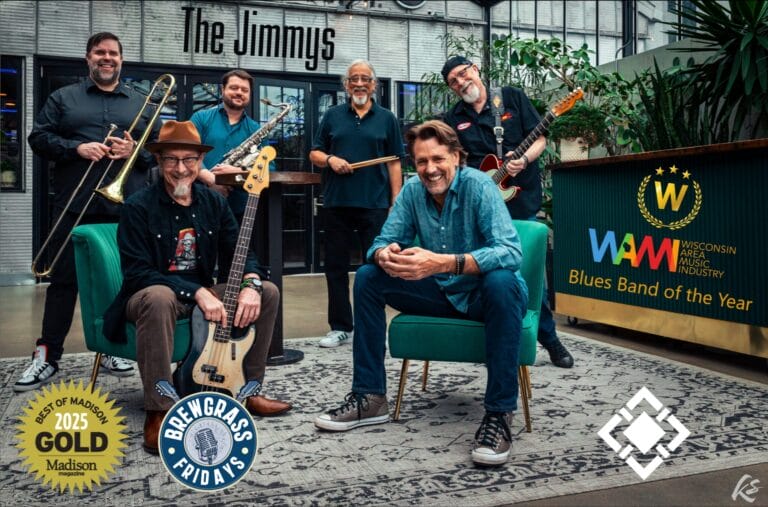 Seven musicians pose together, showcasing instruments in a vibrant indoor setting, with awards displayed prominently.