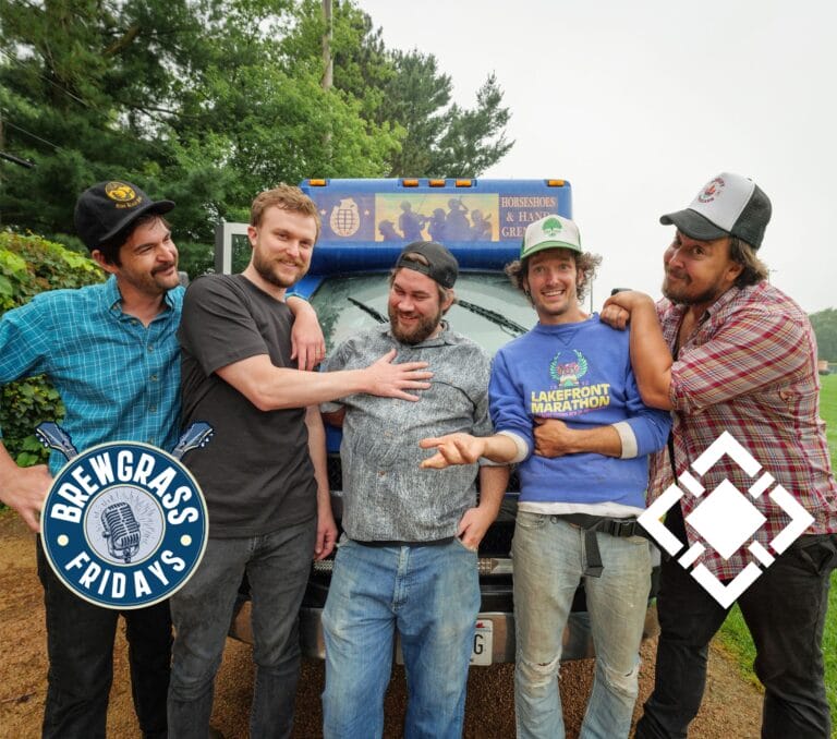 Five men pose playfully in front of a truck, wearing casual clothes and smiling, with greenery in the background.