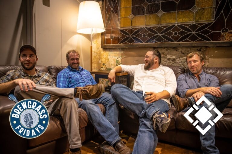 Four men relax on a couch, laughing and chatting, with a lamp and stained glass backdrop enhancing the cozy atmosphere.