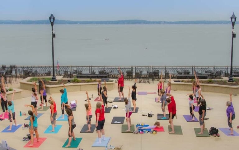 A group of people practicing yoga outdoors by the water, with mats in various colors and a scenic view of the lake.