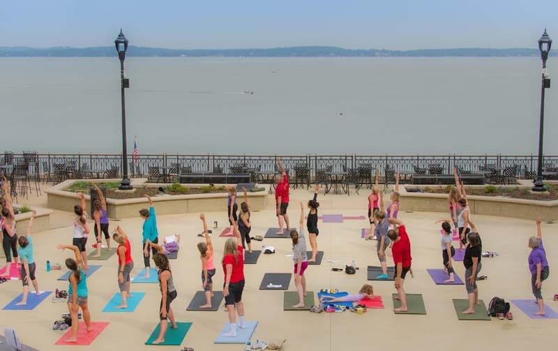 A group of people practicing yoga outdoors by the water, with mats in various colors and a scenic view of the lake.
